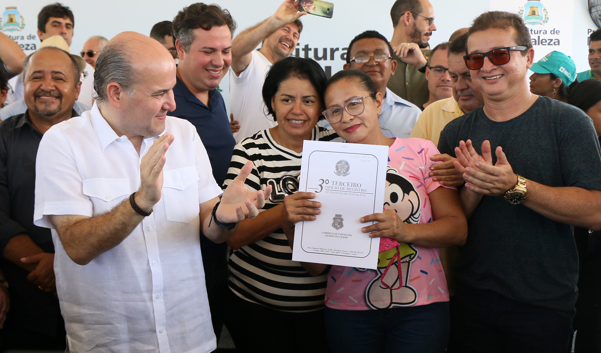 prefeito em pé aplaudindo e pessoas com documentos nas mãos sorrindo para a foto